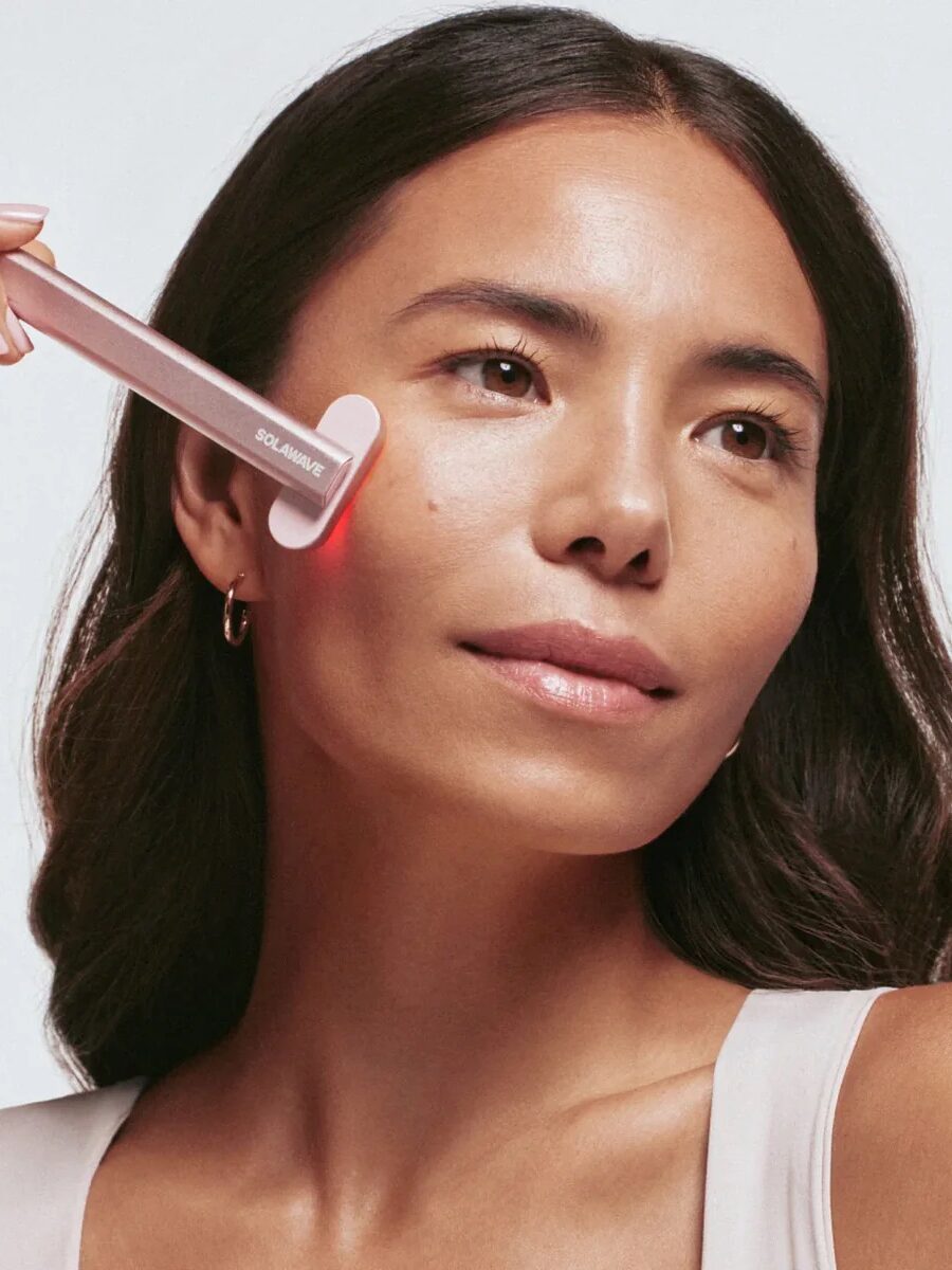 A woman uses a skincare device on her face that emits red light. She is looking slightly upward and smiling softly.