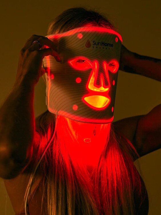 A person wearing a white LED light therapy mask emitting red light, labeled "SunHome." The mask covers the face and neck.