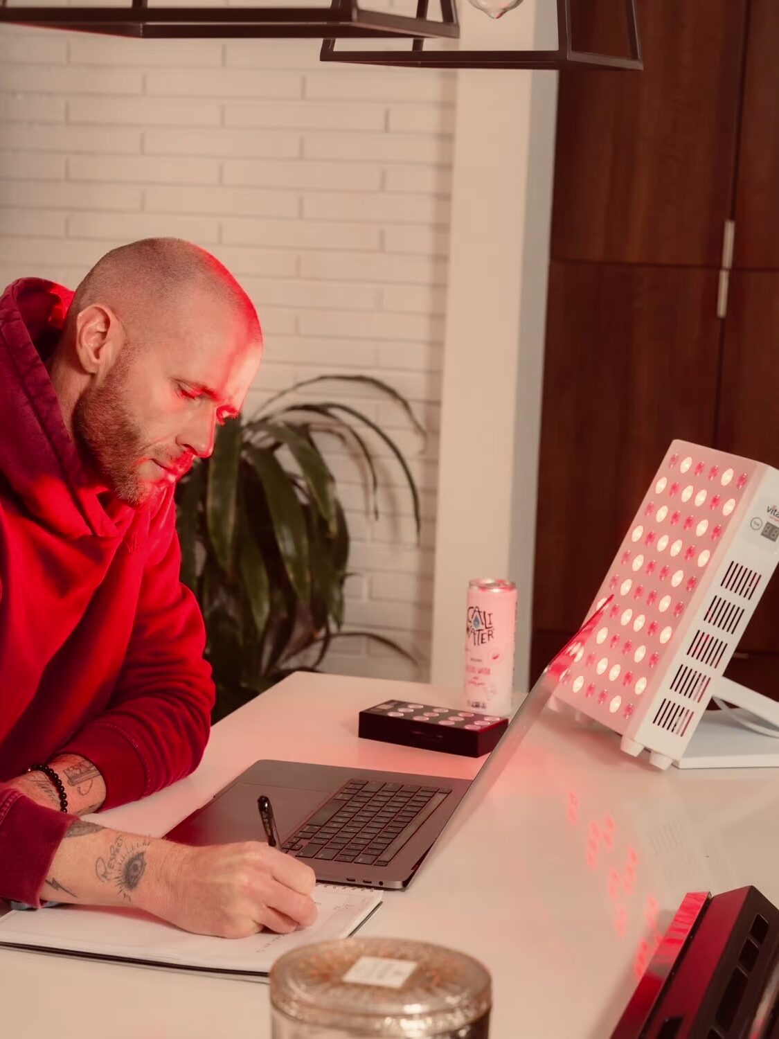 A person in a red hoodie is sitting at a desk, writing in a notebook with a laptop and beverage can nearby, illuminated by a red light.