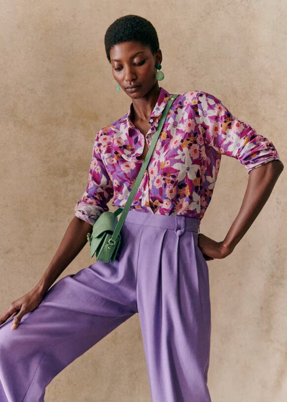 Person wearing a floral-patterned purple shirt, light purple pants, and a green crossbody bag. They are posing against a beige background.