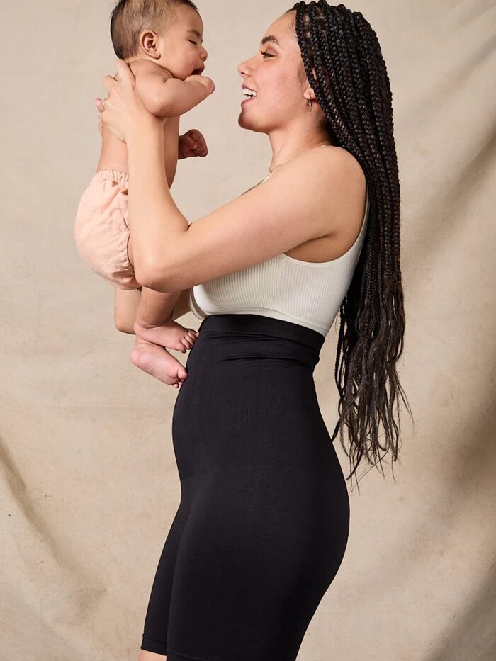 A person with long braided hair, wearing a white top and black shorts, holds a baby dressed in a light-colored diaper against a beige backdrop.