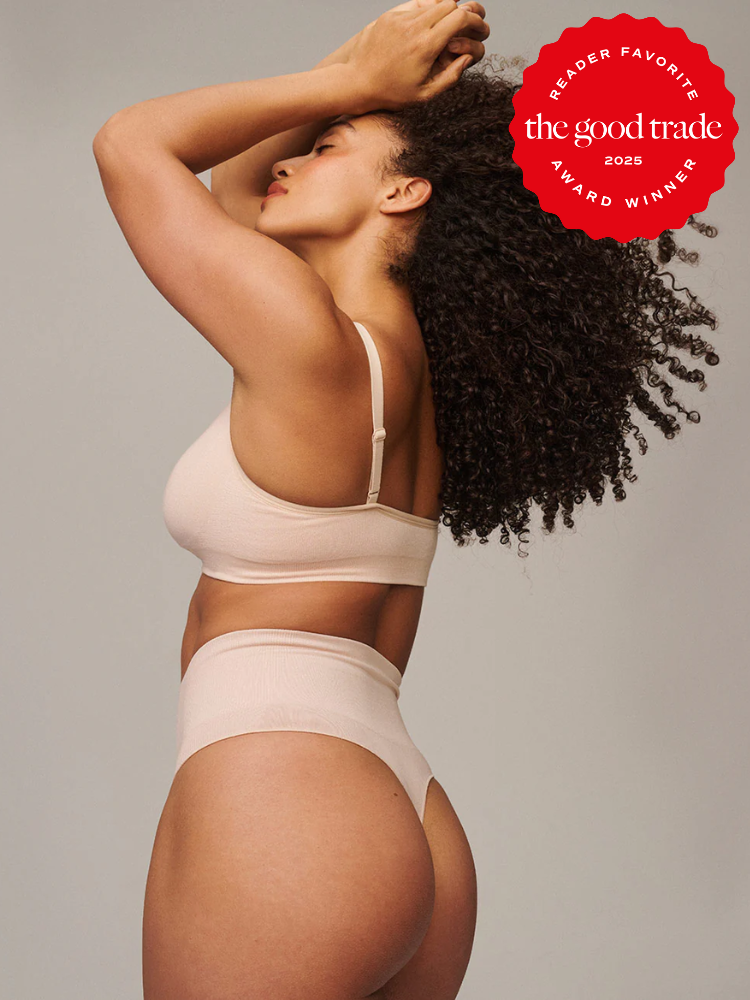 Person wearing beige undergarments poses with arms raised. Curly hair cascades down their back. A red "The Good Trade 2025 Award Winner" badge appears in the top right corner.