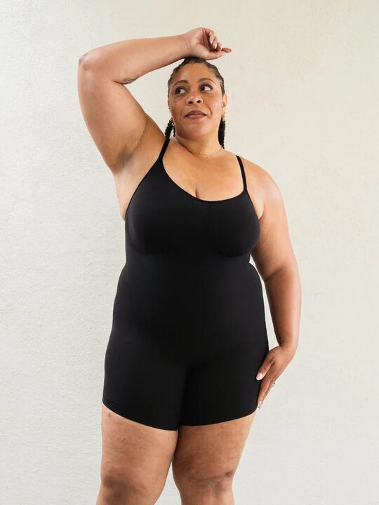 Person wearing a black sleeveless bodysuit stands with one arm raised and the other by their side, posing in front of a plain light-colored background.