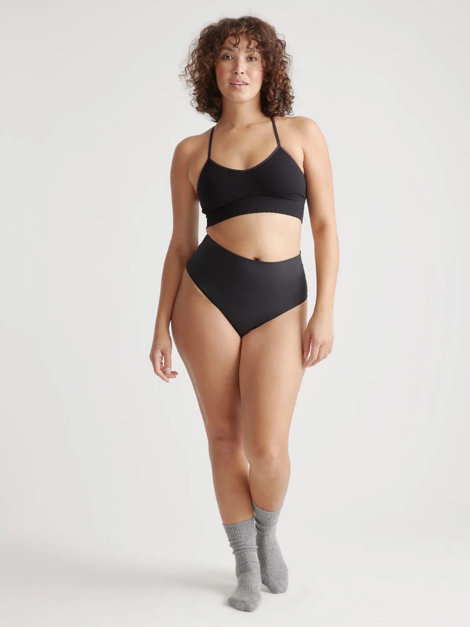 A person stands against a plain background wearing a black bikini top and high-waisted black bikini bottoms, with gray socks. The individual has curly hair and poses with a relaxed expression.