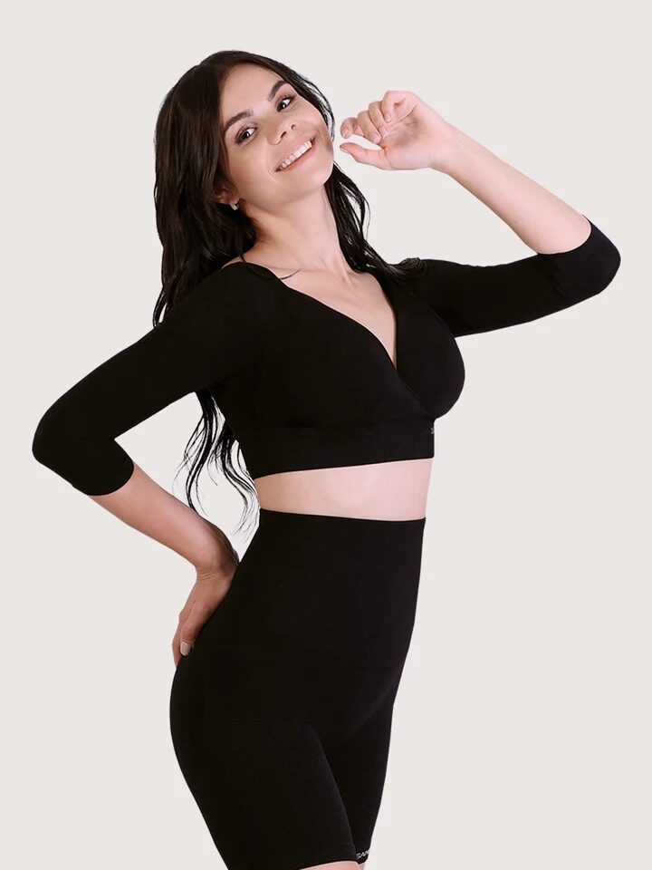 A woman with long dark hair wearing a black, form-fitting long-sleeve top and high-waisted shorts poses with a hand raised near her face, smiling at the camera.