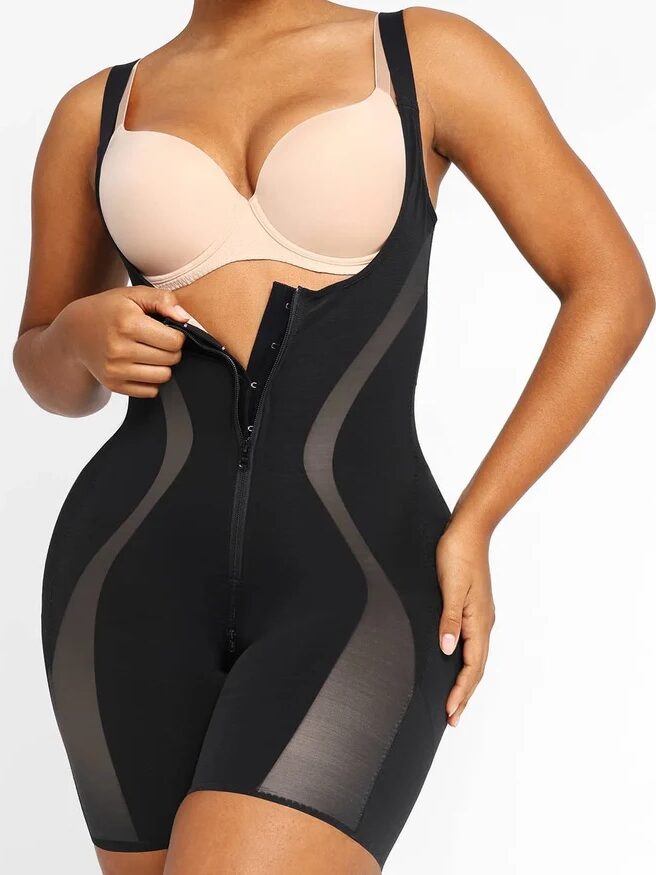 A person is wearing a black body shaper garment with sheer panels, and a beige bra. The body shaper has a front zipper and hook-and-eye closure.