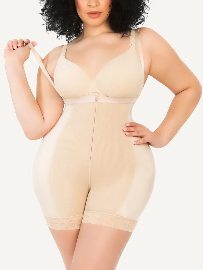 A person wears a beige shapewear bodysuit. They are adjusting the strap on their left shoulder. The bodysuit extends to mid-thigh, featuring detailed hem and front zipper closure.