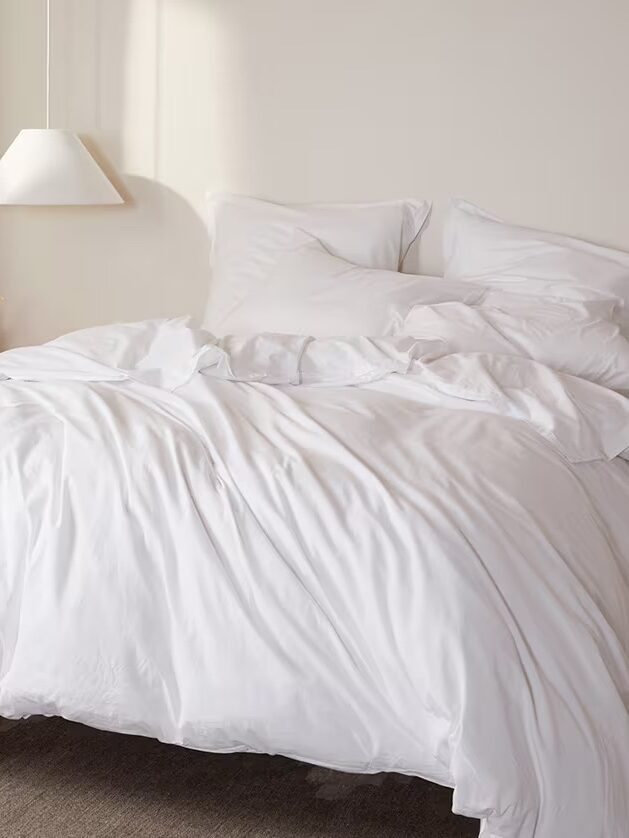 A neatly made bed with white sheets, pillows, and a comforter in a minimalist bedroom with a hanging light fixture and neutral-colored walls.