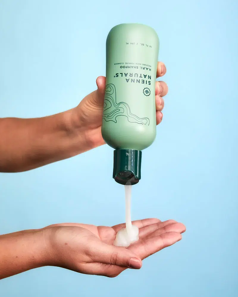 A pair of hands holding a green bottle of shampoo, dispensing a white creamy substance into one hand against a light blue background.