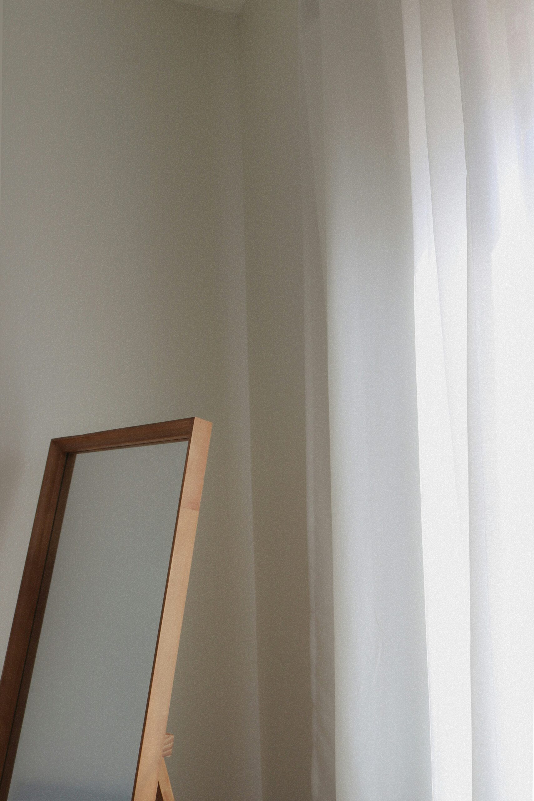 A simple wooden-framed mirror stands adjacent to white sheer curtains in a bright corner of a room.