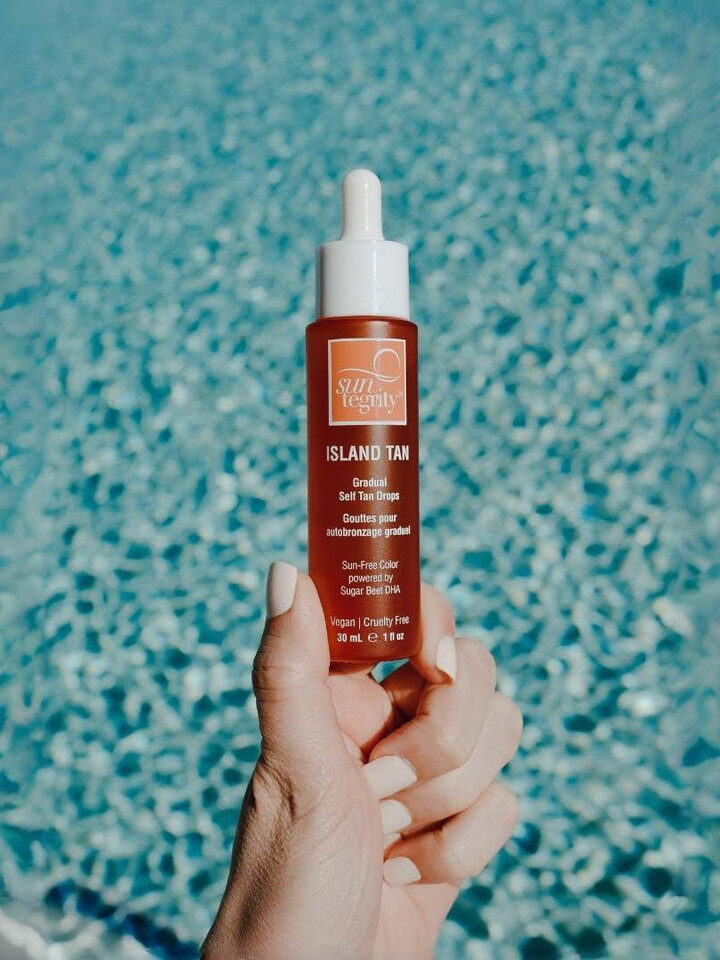 A person holds a bottle of Suntegrity Island Tan in front of a clear blue pool. The product label mentions that it's safe for sensitive skin, vegan, and cruelty-free.