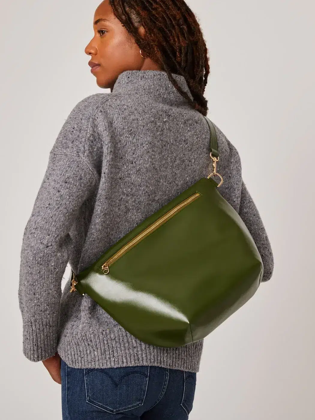 A person wearing a gray sweater and jeans is seen from the side carrying a large green leather shoulder bag with a zipper pocket.