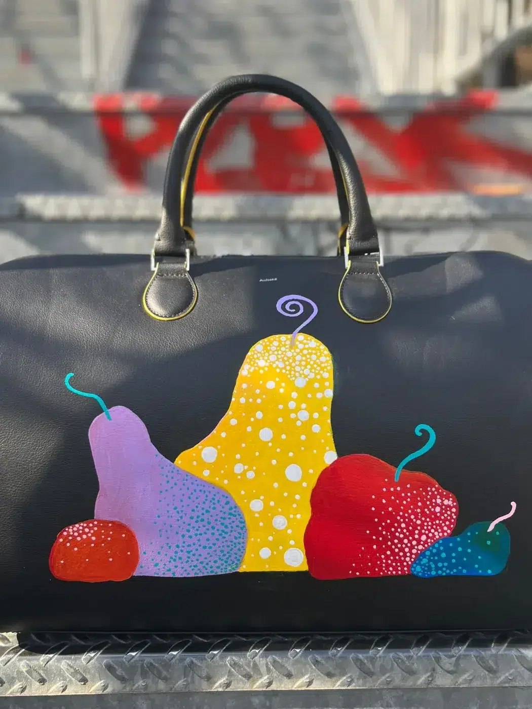 A black handbag with colorful pear-shaped designs in red, yellow, pink, and blue sits on metal stairs with red paint splatters in the background.