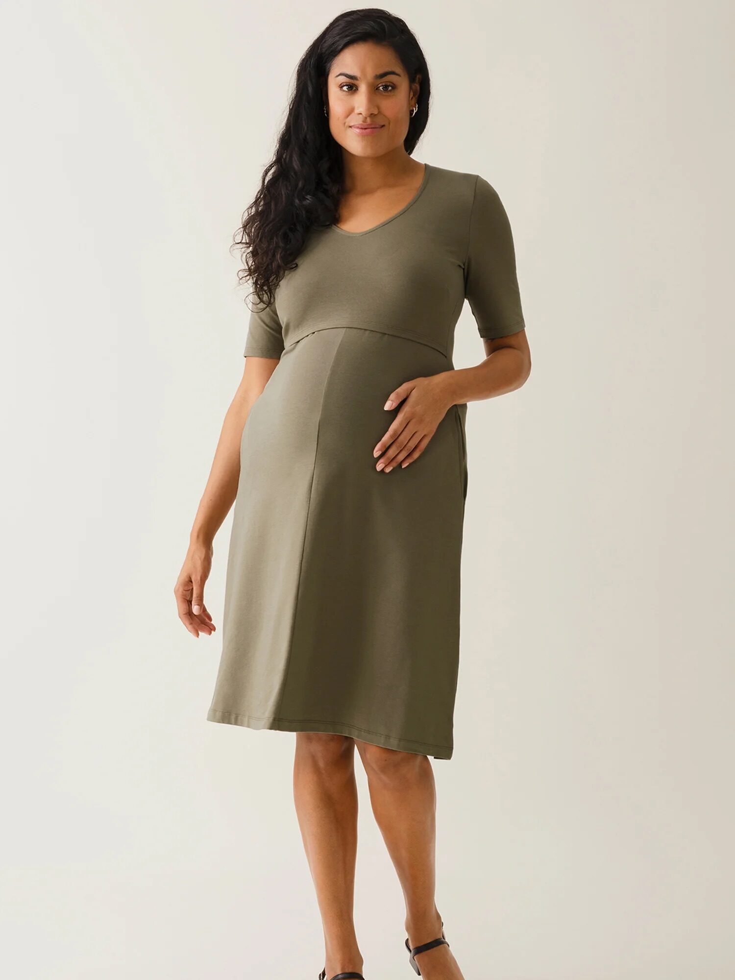 A woman is standing wearing a knee-length, olive green maternity dress with short sleeves. She has one hand on her abdomen and is smiling slightly.