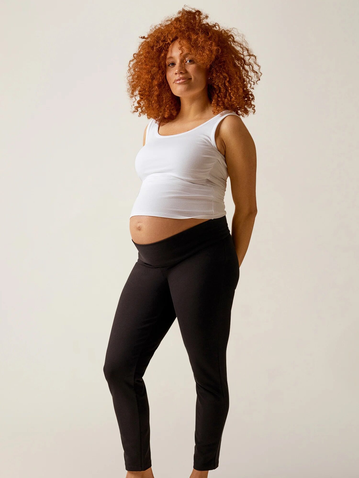 Pregnant person with curly red hair wearing a white tank top and black pants stands barefoot against a plain background.