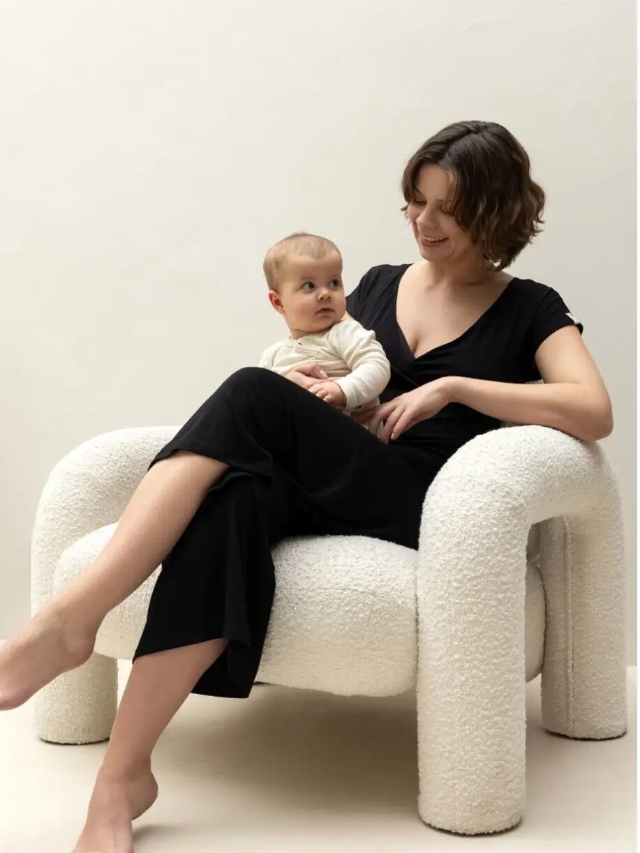 A person sits in a white, plush chair, holding a baby dressed in light-colored clothing. The person is wearing a black outfit and smiling at the child, who is looking up at them.