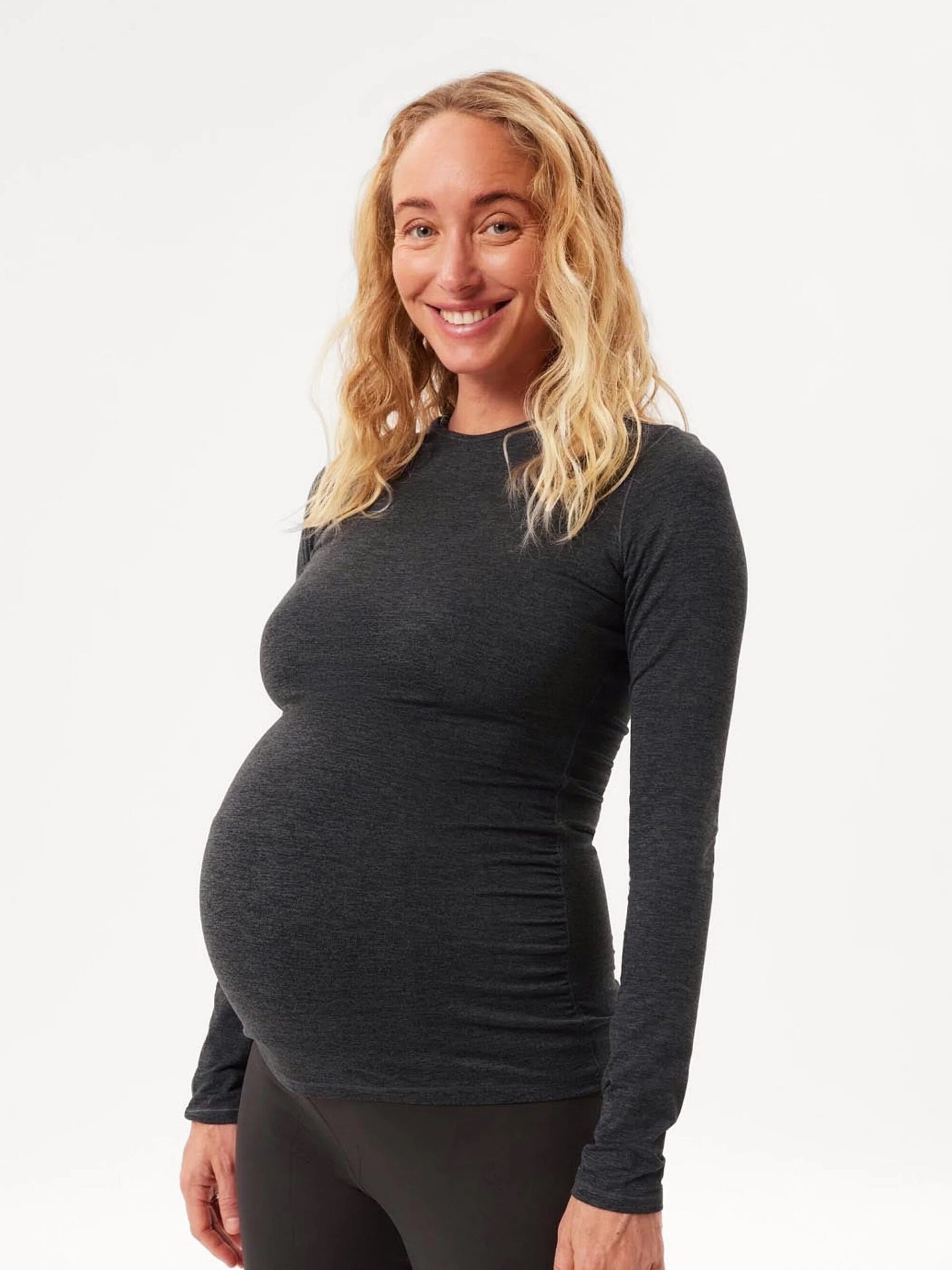 A smiling woman with long blonde hair, wearing a fitted, long-sleeved, dark grey top, and black pants, stands facing the camera. She appears to be pregnant.