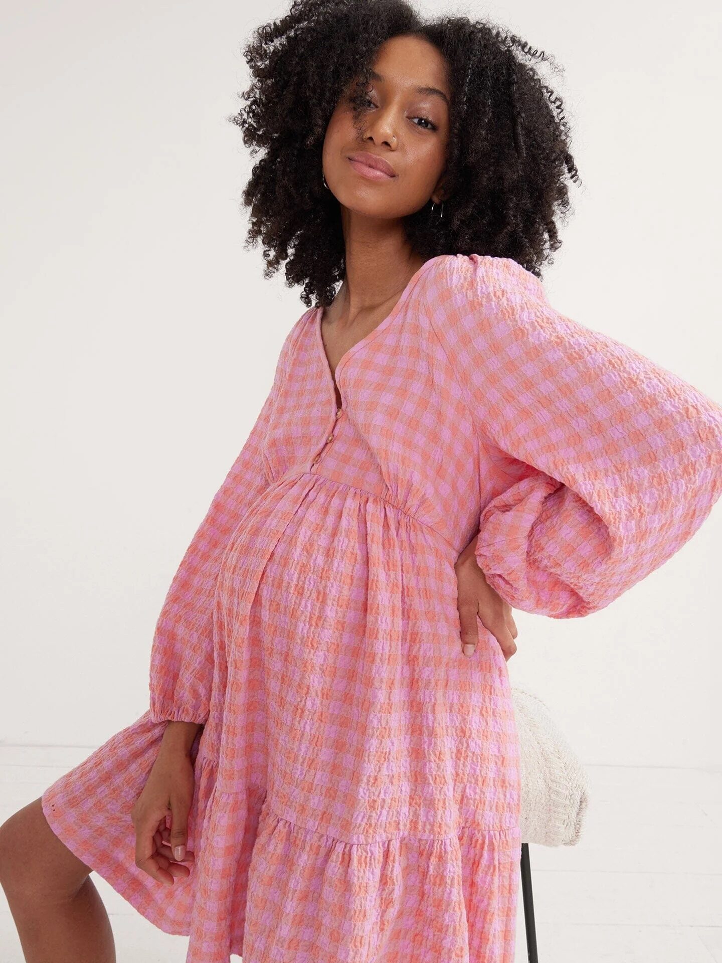 A woman in a pink checkered dress with long sleeves is posing, holding one hand on her hip and the other on her waist, showcasing her pregnant belly.