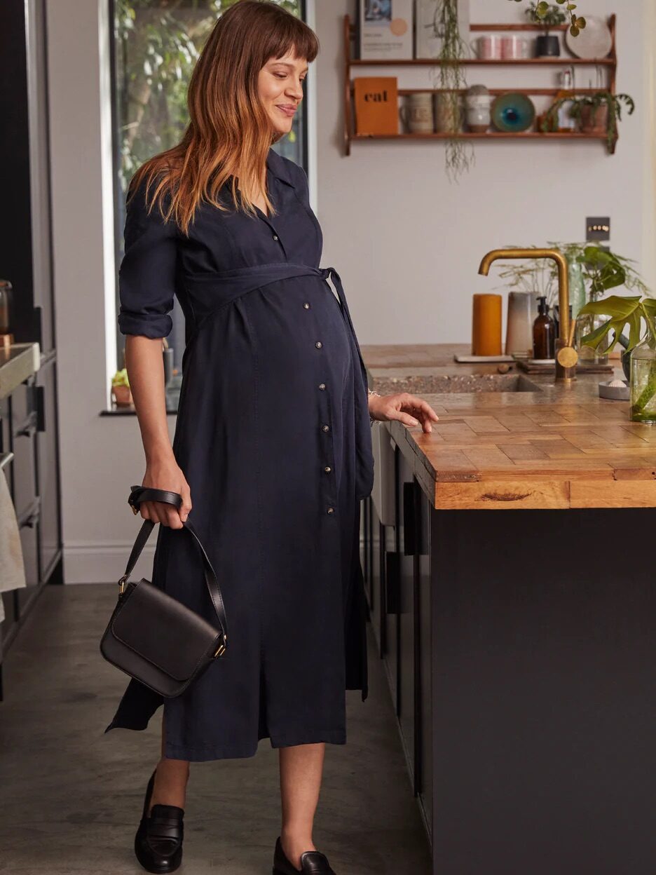 A pregnant woman in a dark dress stands in a modern kitchen holding a black handbag, with one hand resting on a wooden island counter. Shelves with decor items are visible in the background.