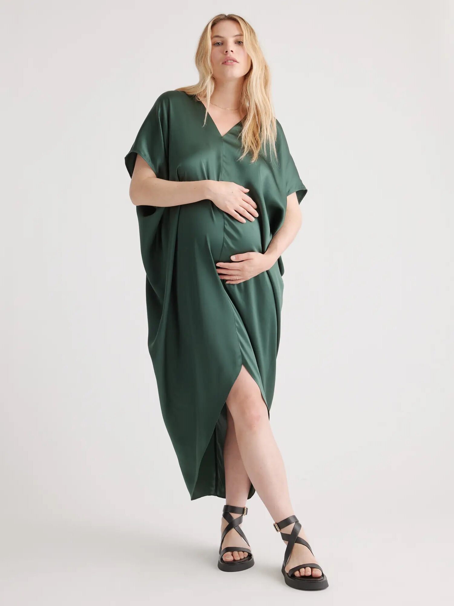 A person wearing a loose, dark green dress and black strappy sandals is posing against a white background. They have long, light-colored hair and are holding their hand on their midsection.