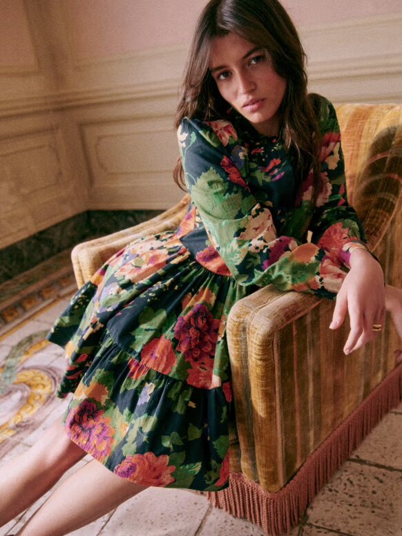 A woman with long dark hair wearing a floral dress sits on an armchair, leaning forward with a neutral expression. The setting is an ornate room with a tiled floor and detailed wall paneling.