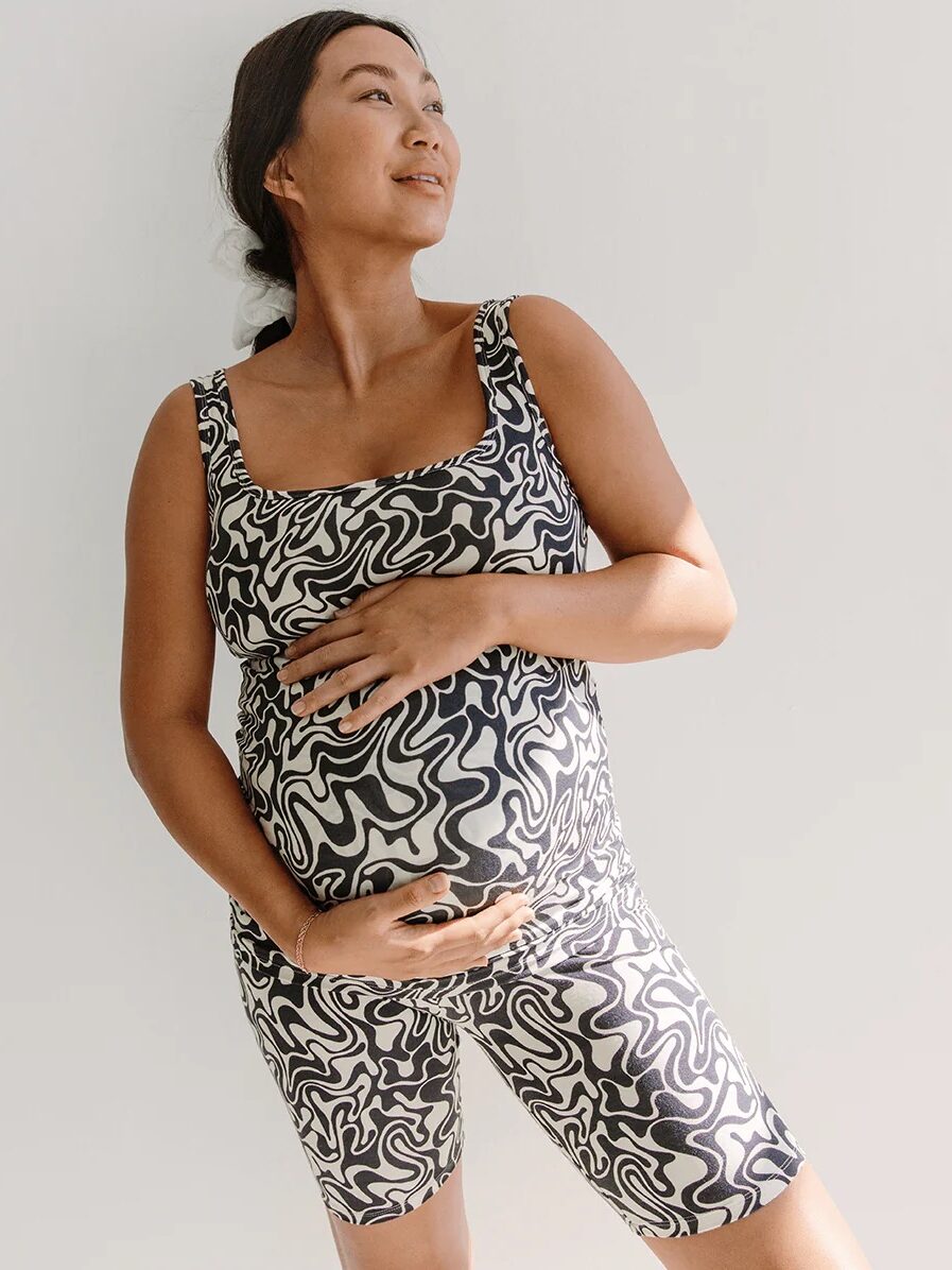 A pregnant woman wearing a matching black and white patterned top and shorts poses with a hand on her belly against a plain background.