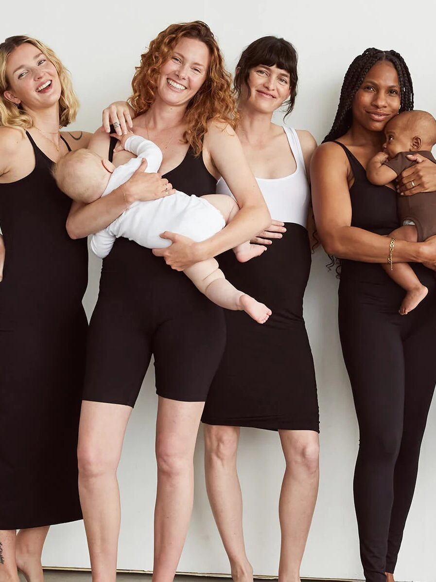 Four women in black and white outfits standing against a plain background, two holding babies.