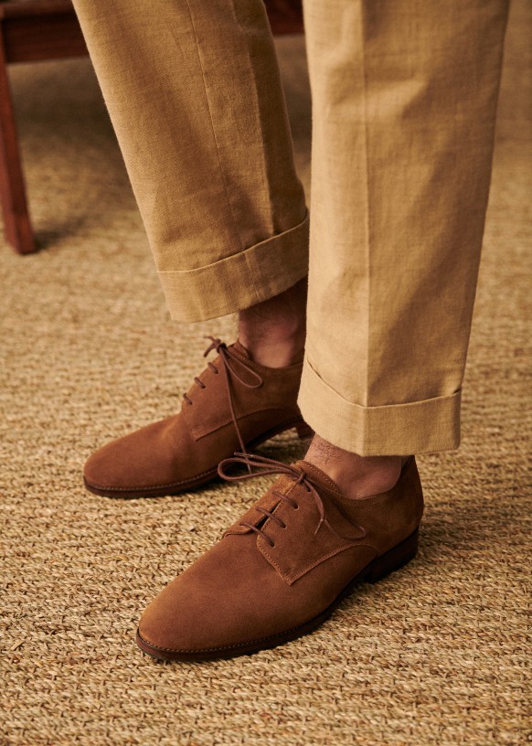 Close-up of a person wearing brown suede lace-up shoes and beige pants with a woven carpet underneath.