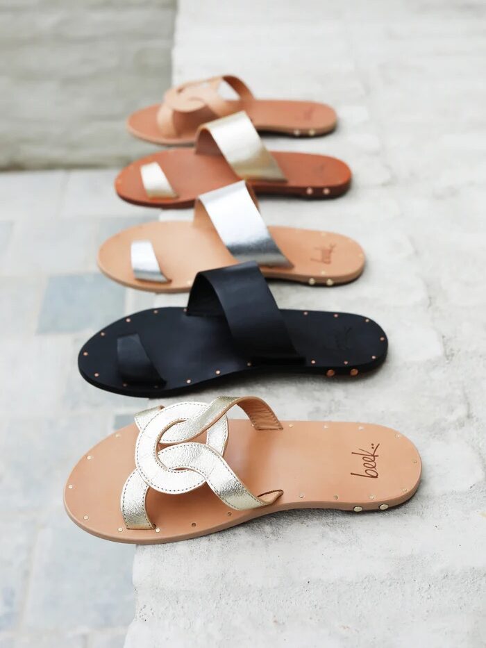 A row of flat sandals in different colors (tan, silver, black, tan, and metallic gold) placed on a light-colored surface. Sandals have varying designs with cross straps and bows.