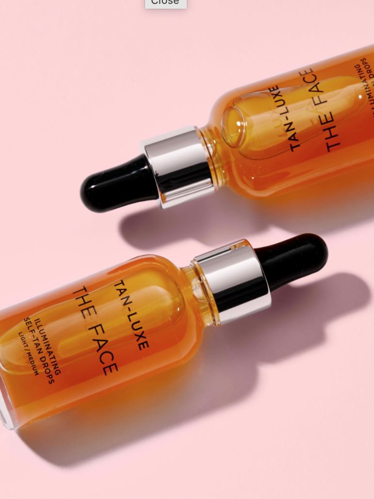 Two amber-colored bottles of Tan-Luxe The Face Illuminating Self-Tan Drops lie on a light pink surface, each with a black dropper cap.