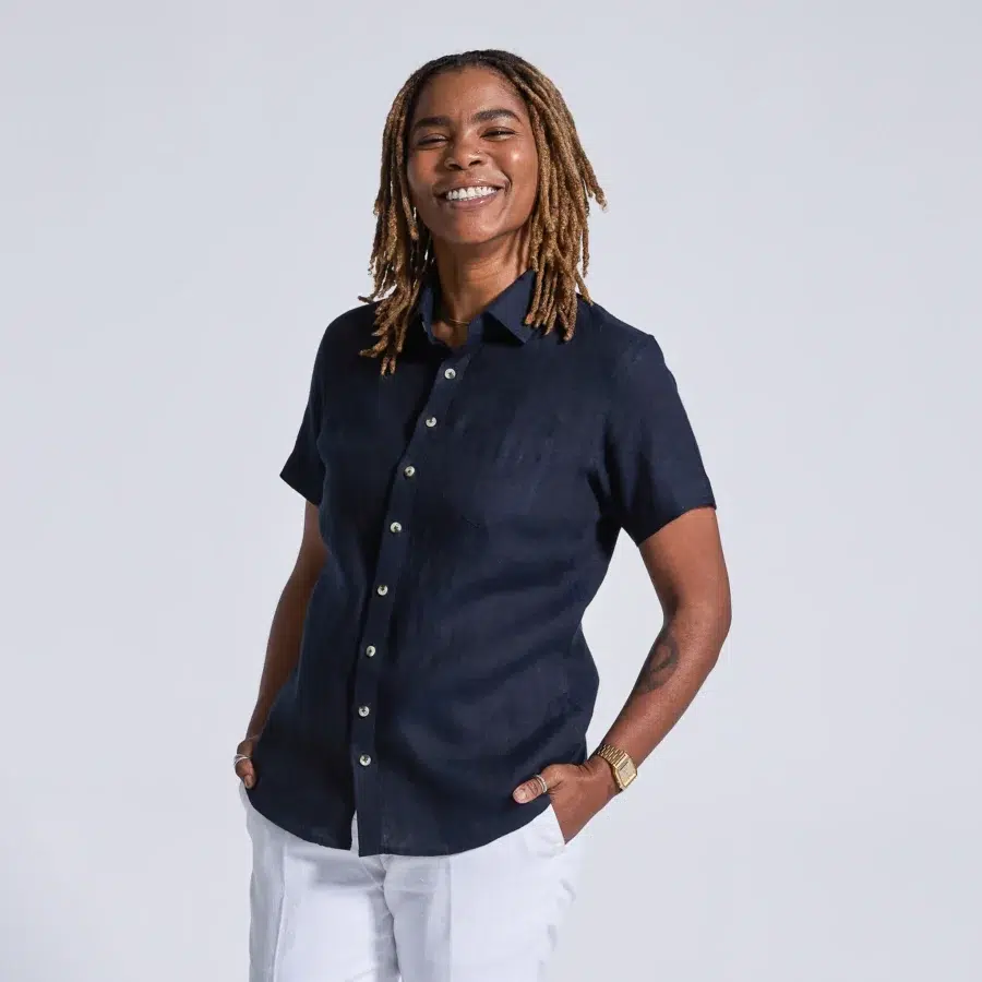 A person with long dreadlocks smiles while standing with hands in pockets, wearing a dark short-sleeve button-up shirt and light-colored pants.