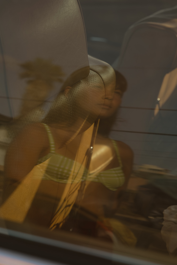 Woman in a green bikini top is seen through a car window with reflections of a palm tree and power lines.