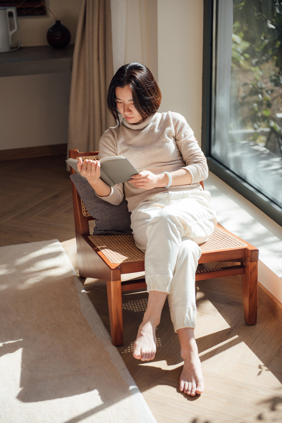 A person sits in a sunlit room reading a tablet on a wooden chair near a window.