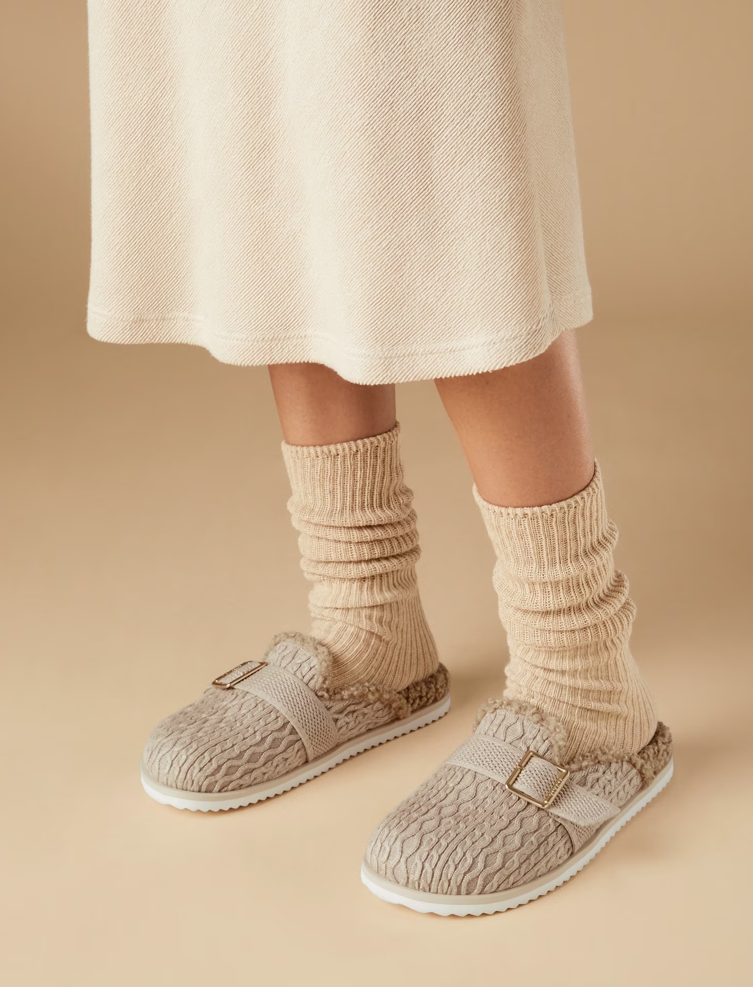 Person wearing a beige knitted skirt, slouchy light brown socks, and beige textured slip-on shoes with a buckle.