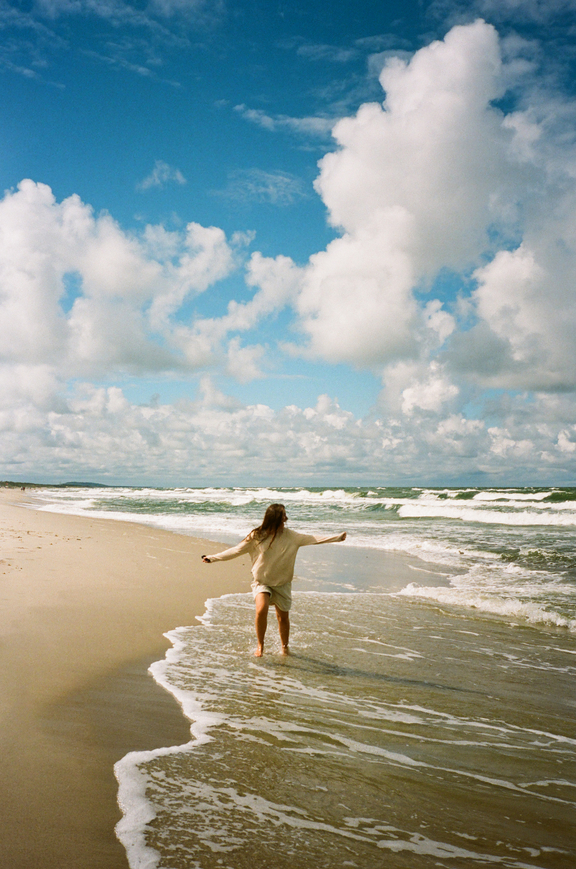 A person walks along a sandy beach with foamy waves under a partly cloudy sky.