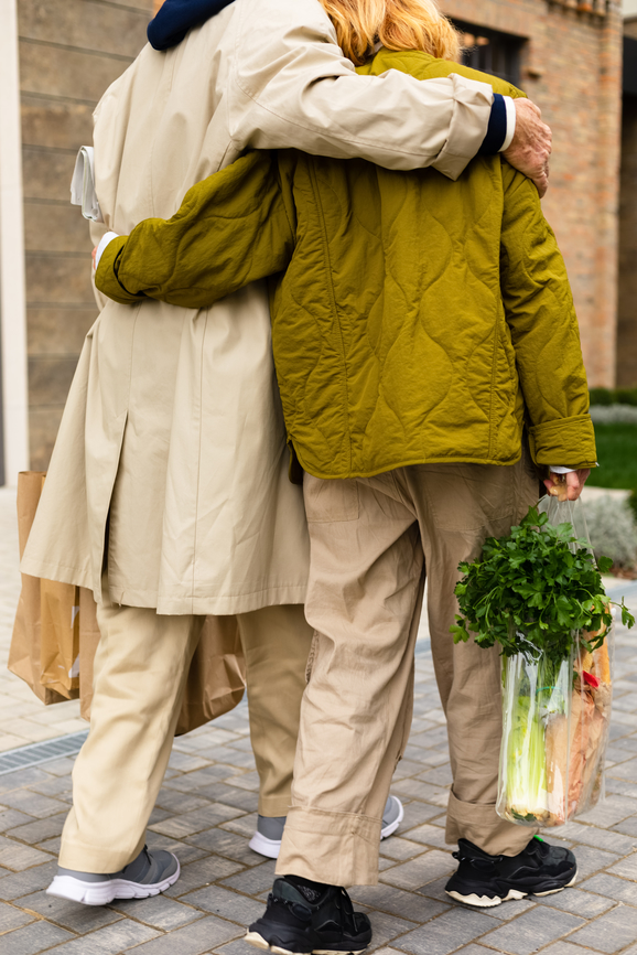 Two people with arms around each other walk on a paved sidewalk, carrying bags of groceries. One wears a green jacket, and the other wears a beige coat.