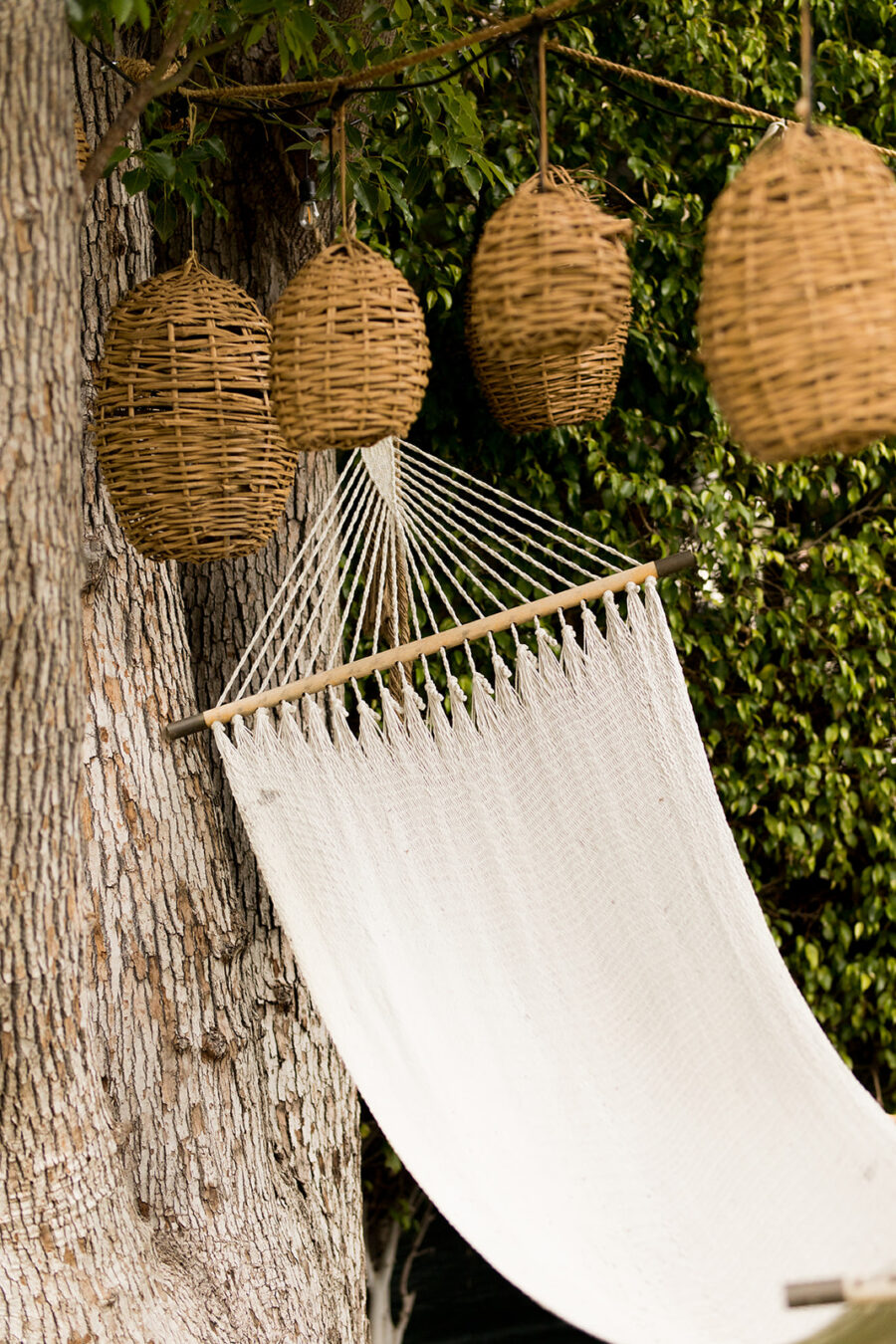 A white hammock is hanging between a tree and large woven lanterns, surrounded by greenery.