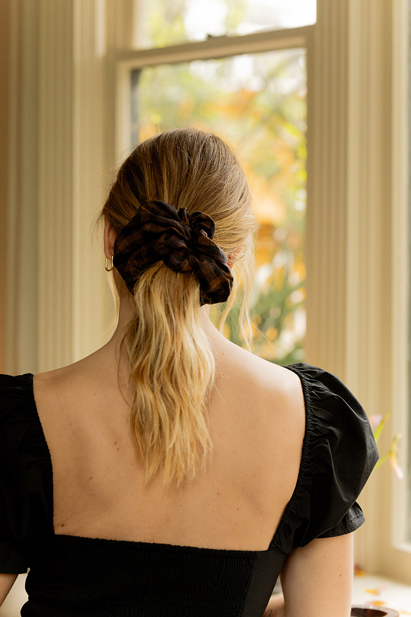 A person with blond hair is seen from the back, wearing a black top with puffed sleeves and a brown plaid scrunchie in their hair, standing in front of a window.