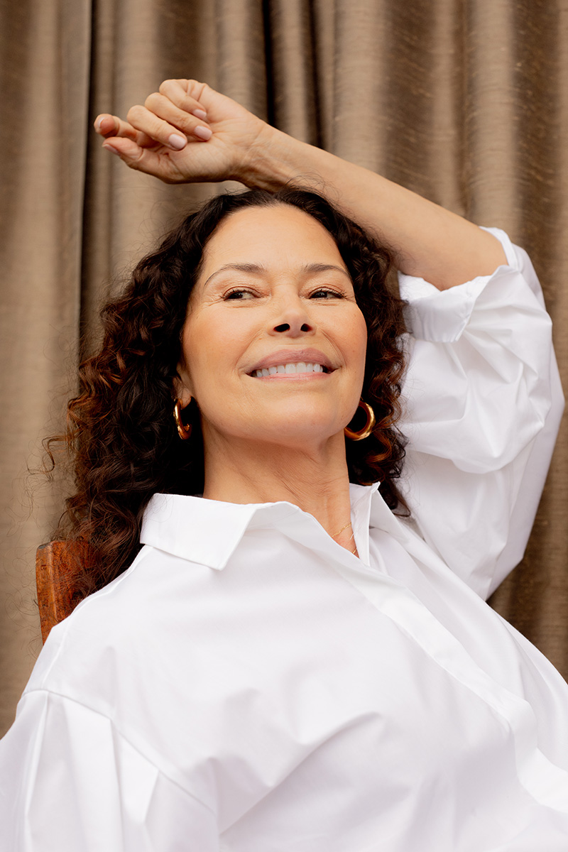 A smiling person with curly hair poses in front of a brown curtain, wearing a white shirt and hoop earrings, with one arm raised overhead.