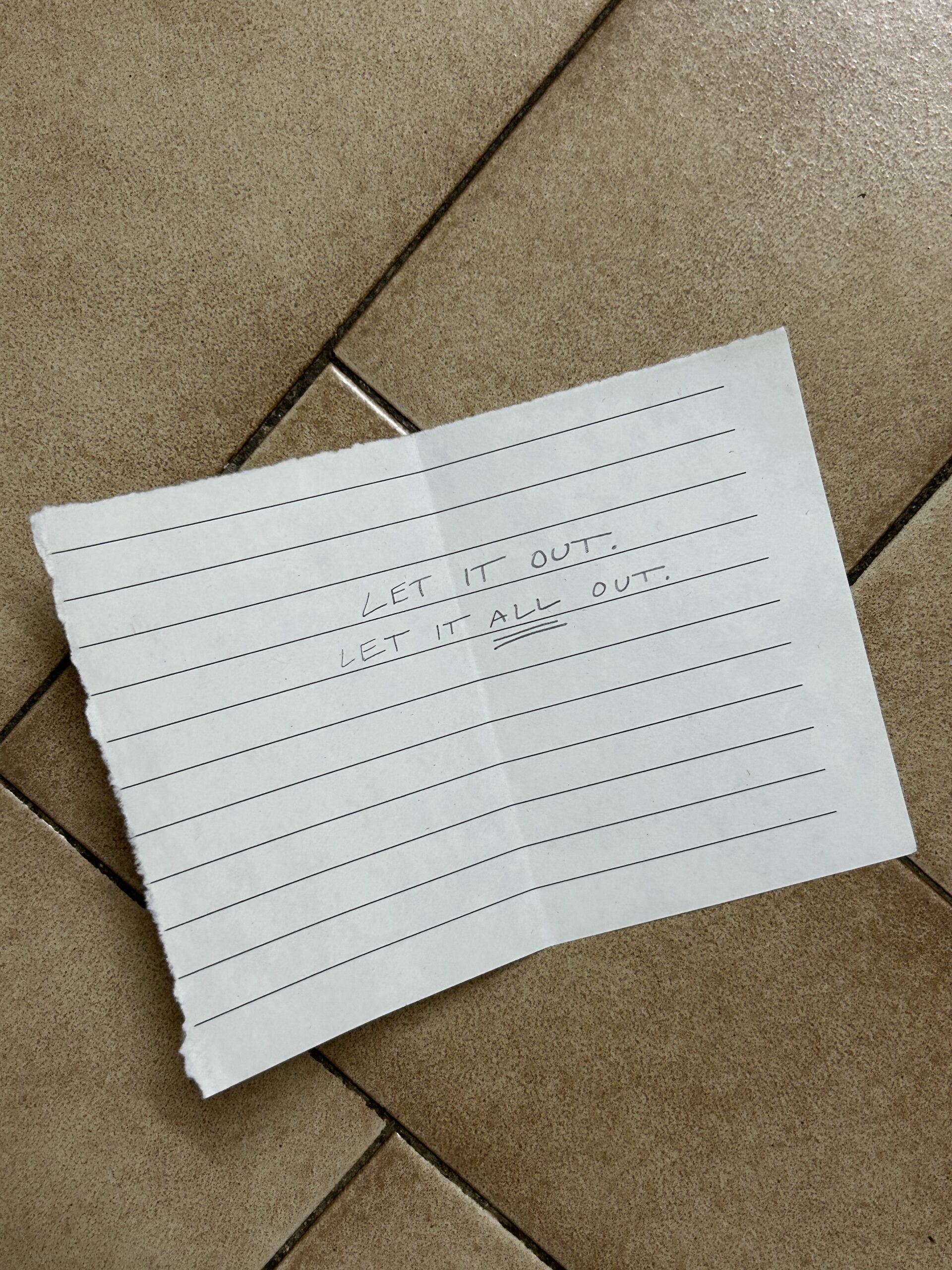 A torn sheet of lined paper on a tiled floor with the handwritten message "LET IT OUT. LET IT ALL OUT.