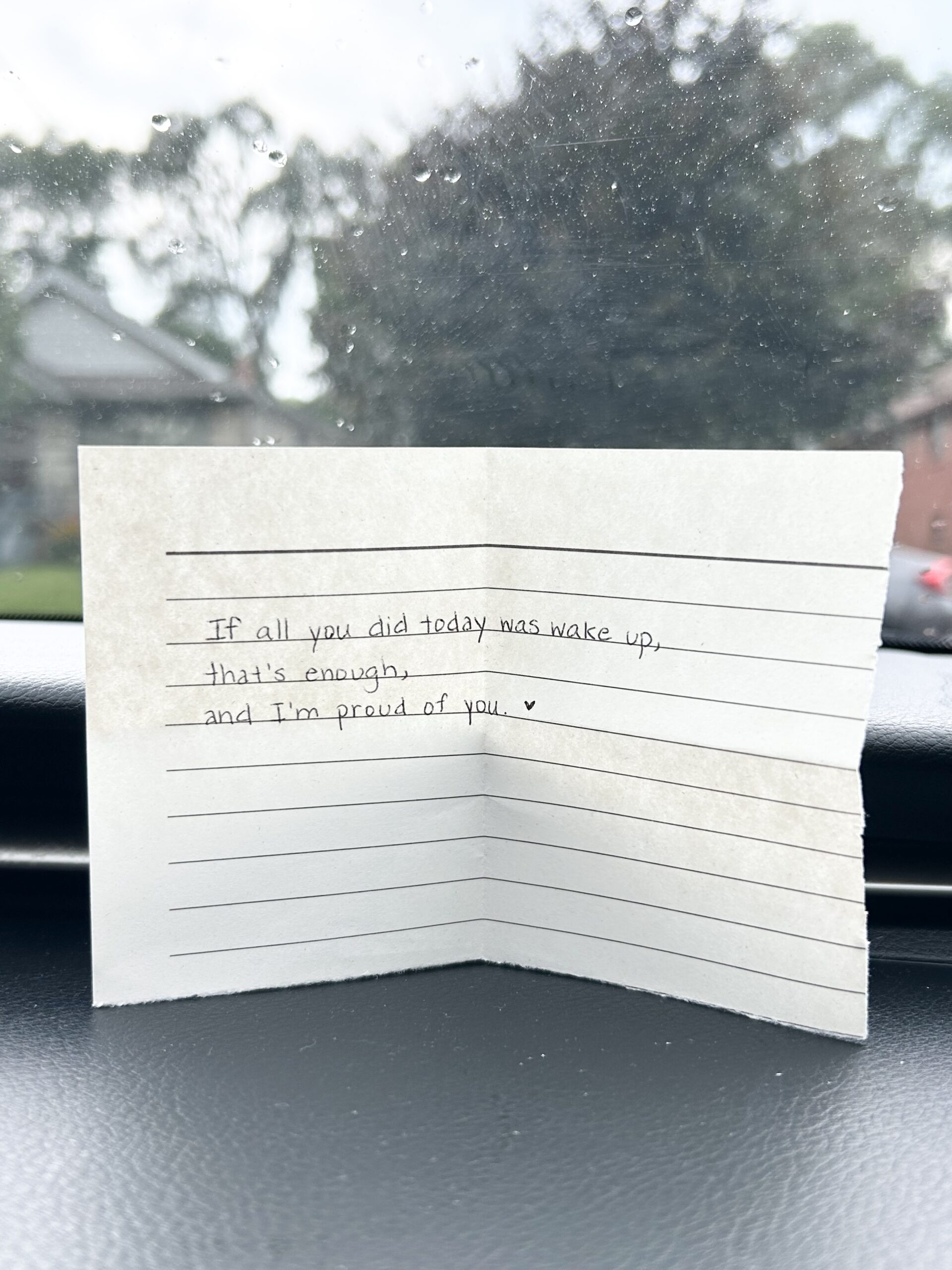 A handwritten note on lined paper reads, "If all you did today was wake up, that's enough, and I'm proud of you." The note rests on a car dashboard with a blurry view of trees and houses outside.