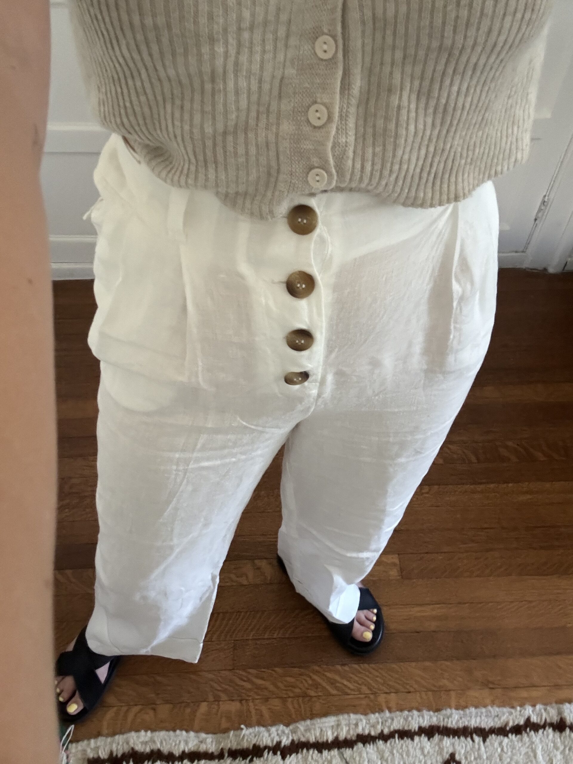 Person wearing white linen pants with a button-down front and a beige knitted top, standing on a wooden floor.