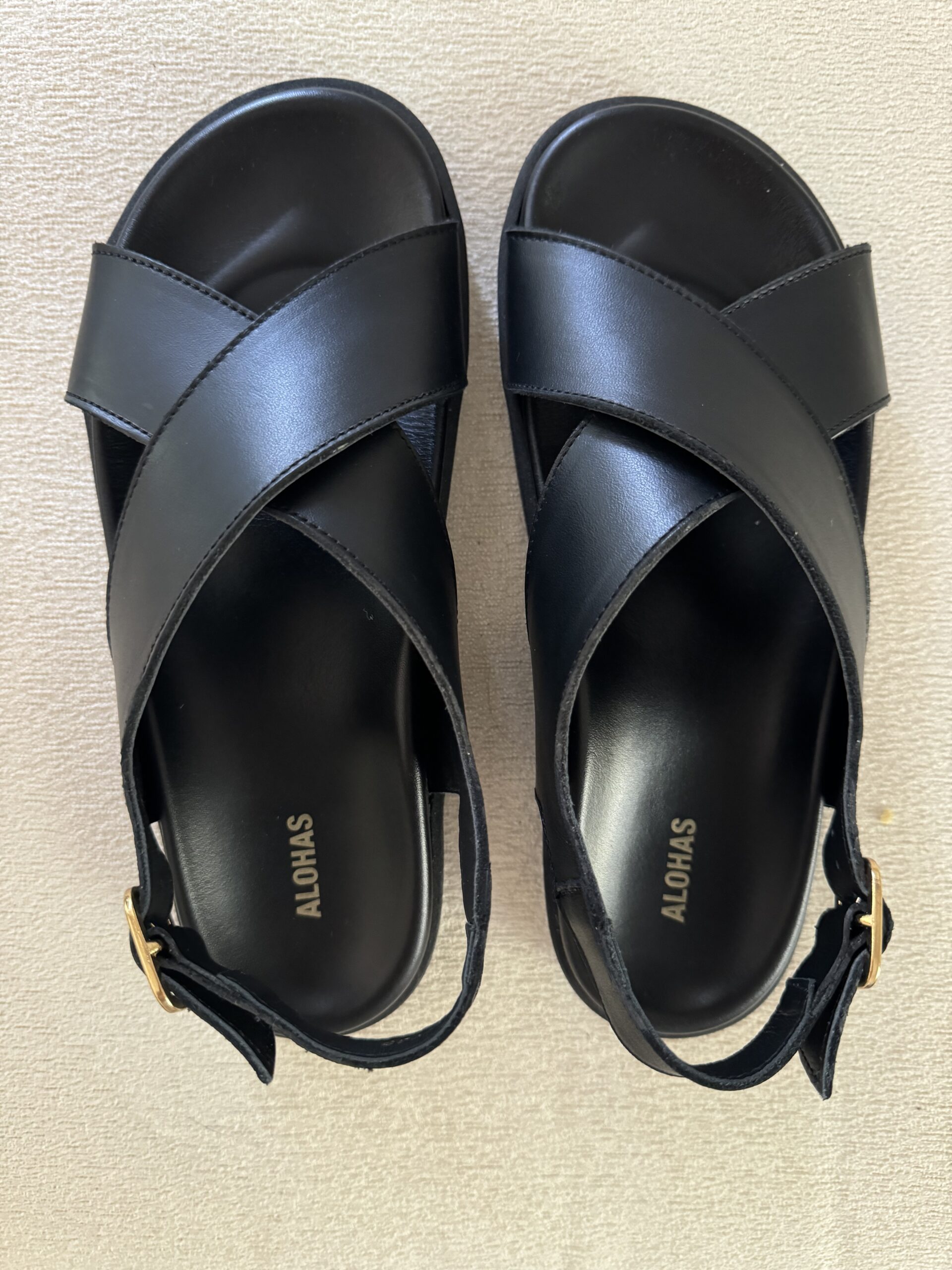 A pair of black, strappy sandals with "ALOHAS" branding on the insole, featuring gold buckles on the ankle straps, placed on a light-colored surface.