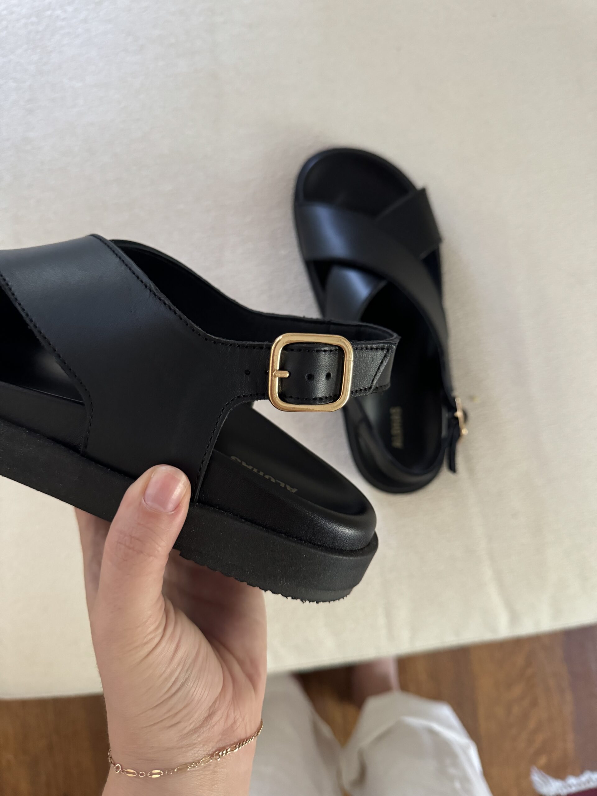 A hand is holding a black sandal with a gold buckle, while another black sandal is placed on a light-colored surface in the background.