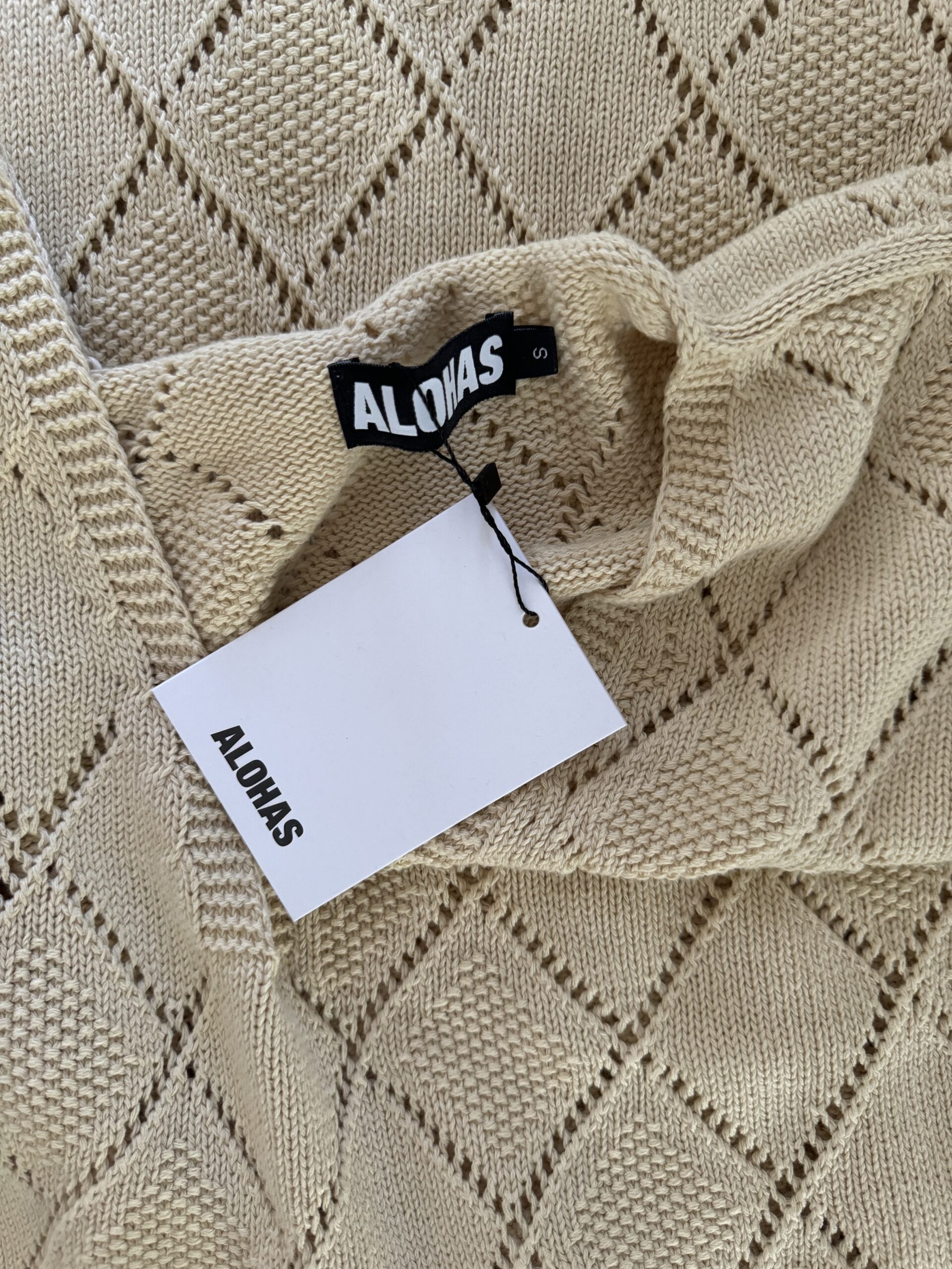 Close-up image of a beige knit sweater with a black "ALOJAS" label and a white tag attached.