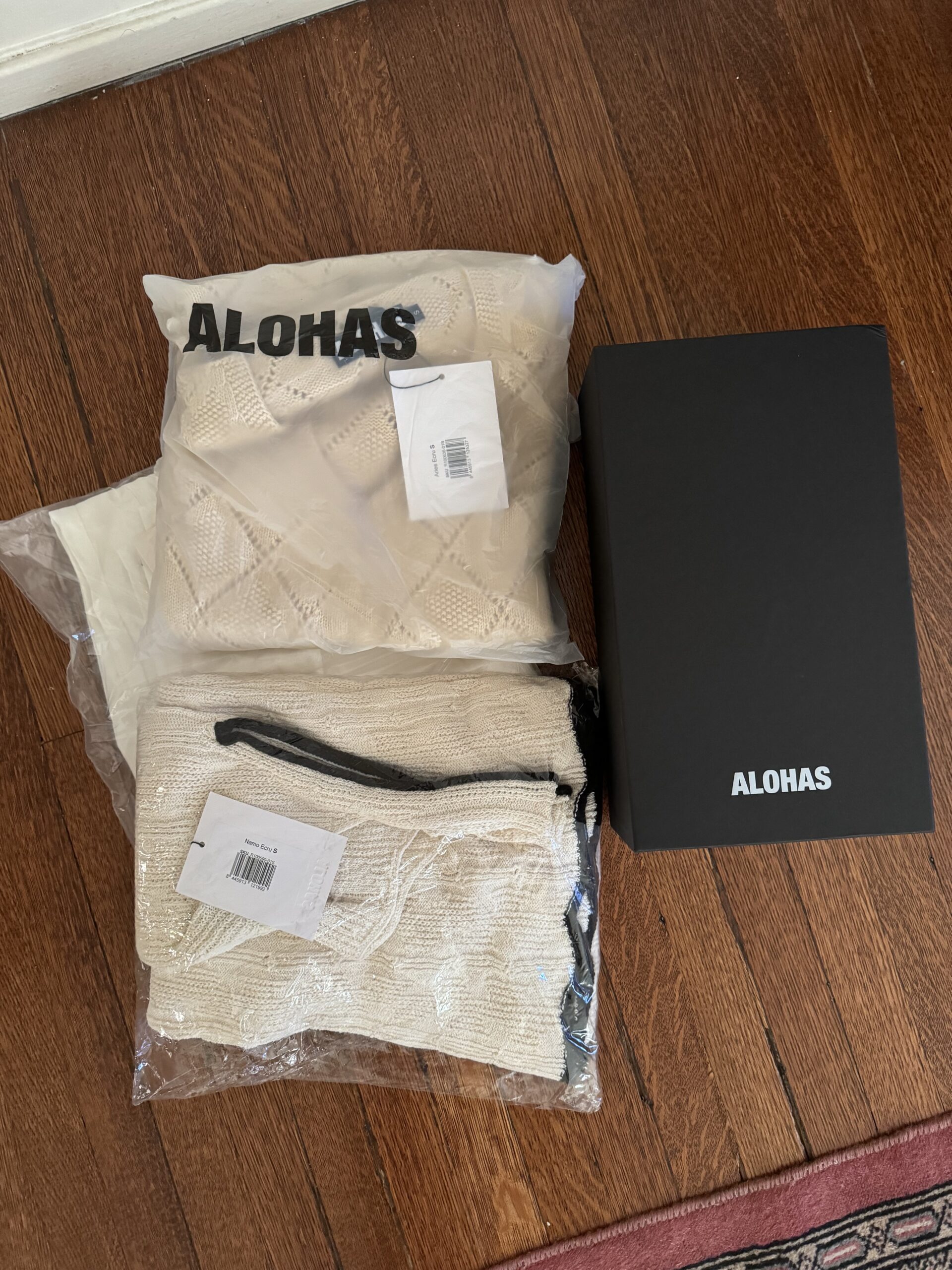 Three packaging bags with "Alohas" printed on them and a black shoe box labeled "Alohas" placed on a wooden floor.