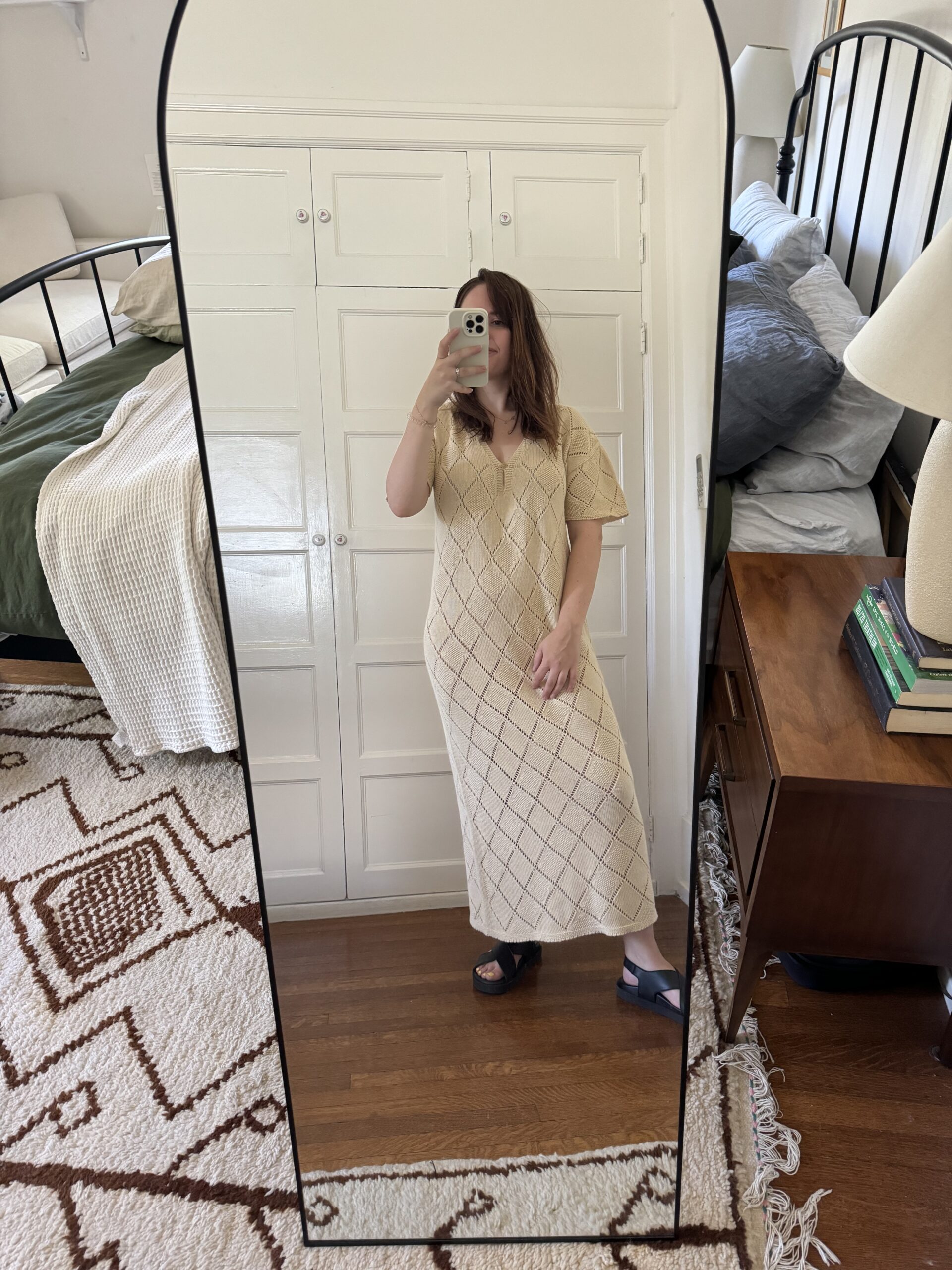 A person stands in front of a full-length mirror wearing a long beige dress and black sandals, holding a phone with one hand. The bedroom in the background has a bed, side table with books, and a lamp.