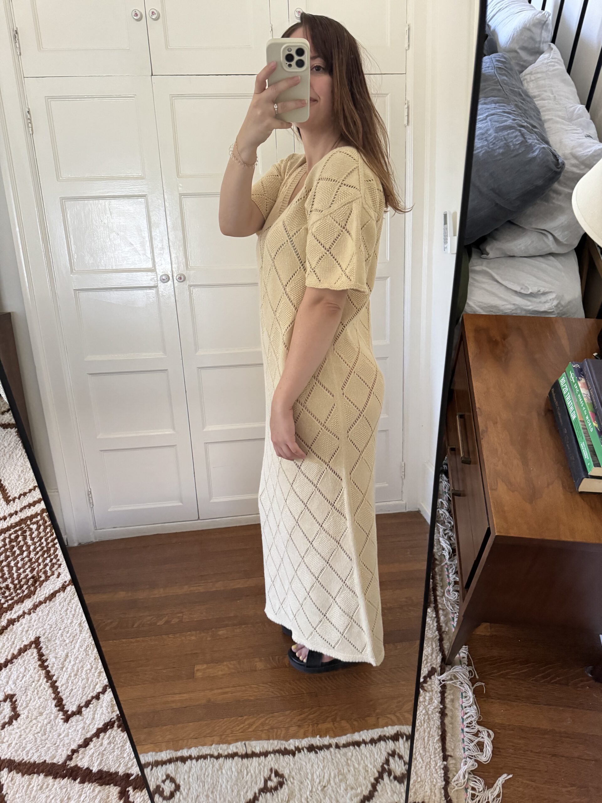 A person in a beige, patterned, ankle-length dress takes a mirror selfie in a room with white cabinets and a wooden nightstand.