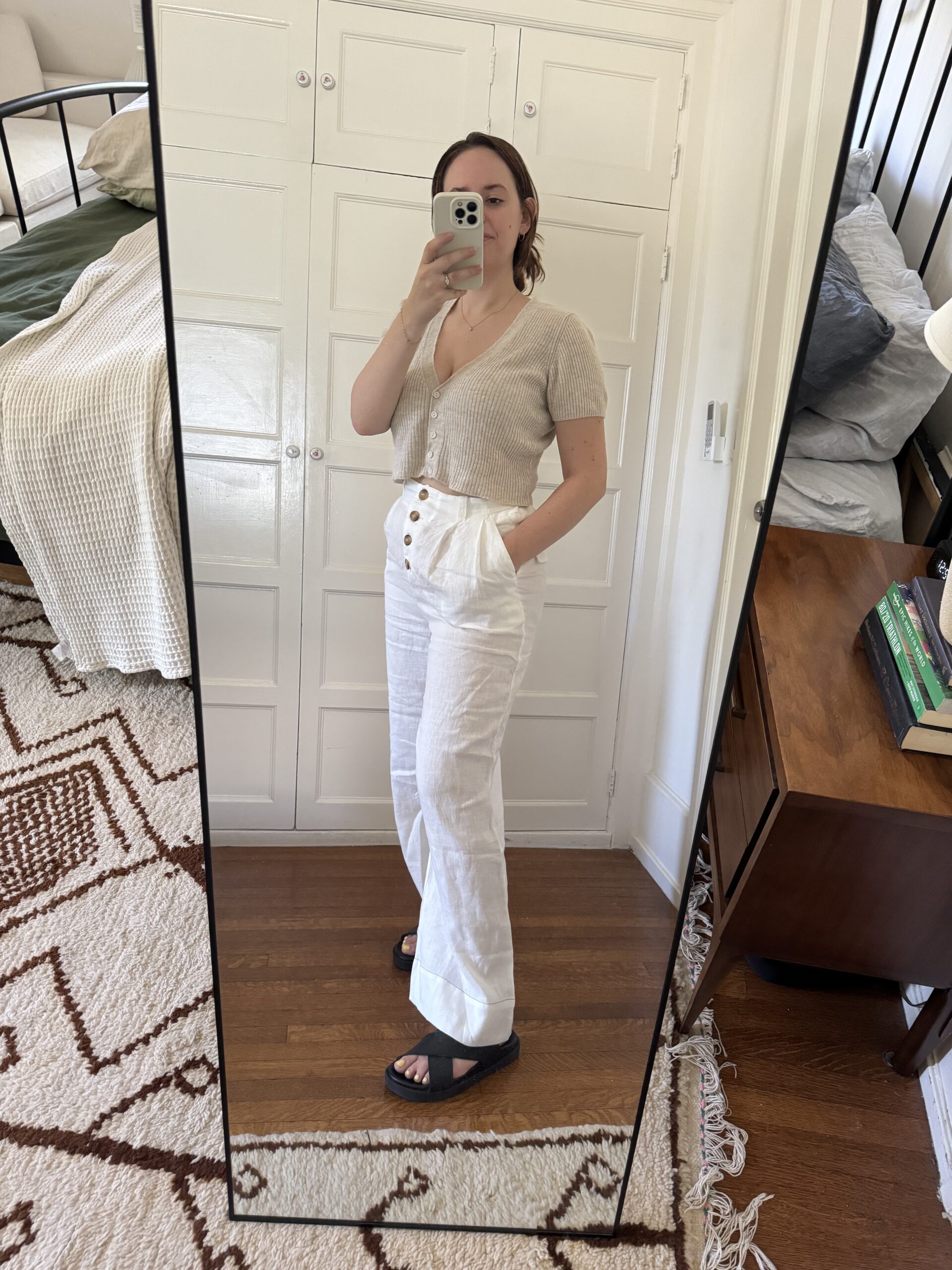 A person takes a mirror selfie wearing a beige top, white high-waisted pants, and black sandals, standing in a room with a bed and a wooden dresser in the background.