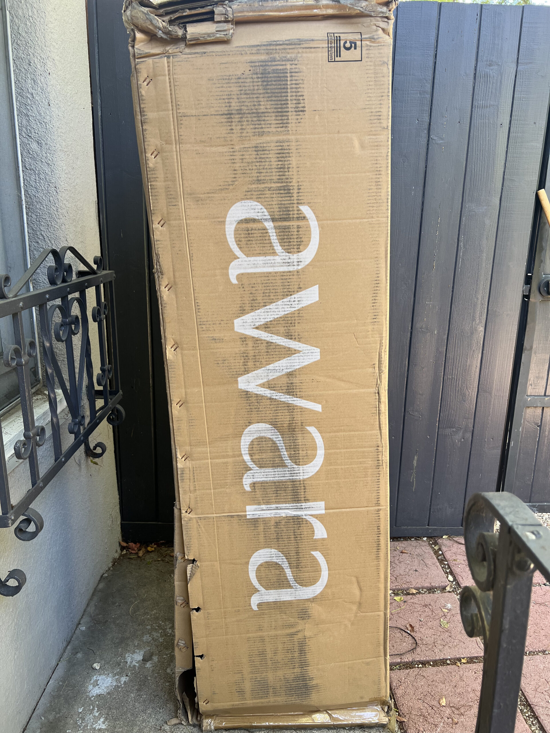 A large brown cardboard box with the word "awara" printed in white lies against a dark wooden fence and a black metal railing. The box appears slightly damaged.